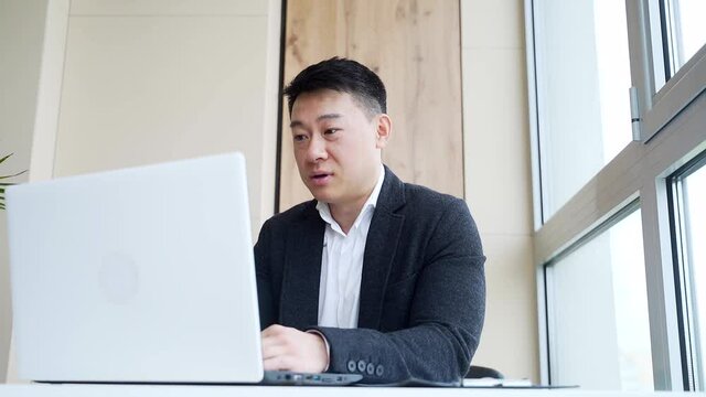 young asian business man sitting in the office talking online via webcam, looking at laptop. Male in formal suit, an employee or manager waving his hand, happy talking on video call indoors. Close up