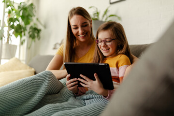 Happy mom or nanny and little girl child rest on sofa watch video on tablet.