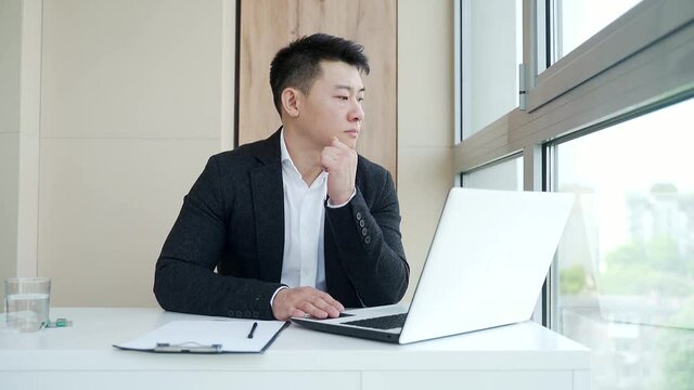 close-up portrait asian pensive young office worker man at workplace. face a serious male business manager in formal suit thinking about solving a problem. Businessman at desk with computer or laptop