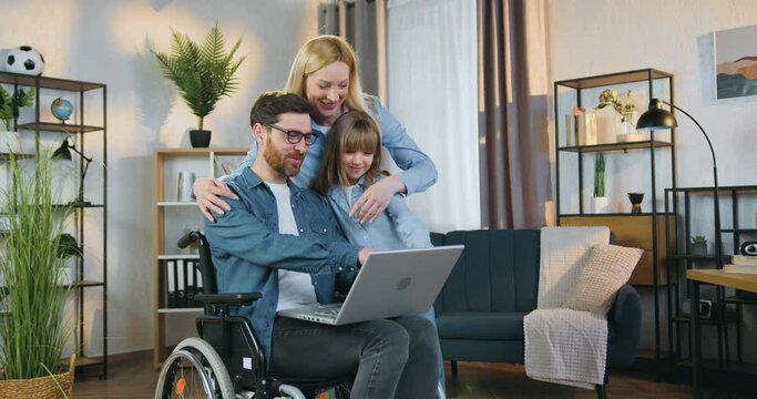 Attractive smiling adult handicapped man in wheelchair applying laptop together with his 10-aged cute daughter and pretty joyful wife at home