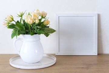 Square wooden frame mockup on a vintage bench, table. Modern white ceramic vase with white roses. White wall background. Scandinavian interior.
