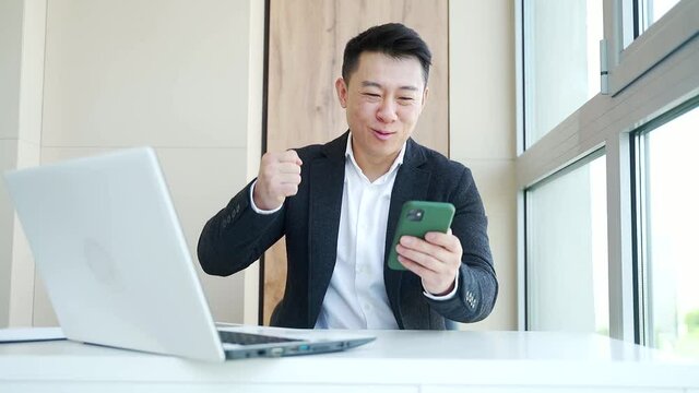 Happy young Asian business man in office looking at mobile phone, with emotion winner or win, financial stock sports betting. Male joyfully exclaims playing game. Excited overjoyed celebrating success