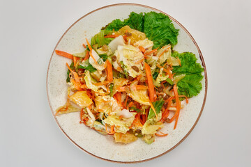 Salad of fresh Peking cabbage, carrots, orange, celery, yellow raisins, seasoned with vegetable oil with a leaf of green lettuce in a round white plate on a white background.