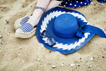 Women's feet in striped sandals, blue hat and sunglasses on the sand.