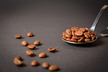 Side view of Roasted coffee beans served in silver vintage stainless steel spoon with scattered beans on dark background. High quality organic aroma seeds. Copy space text image
