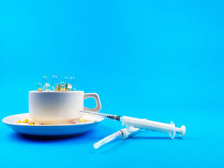 A variety of pills in a coffee cup, syringes next to a blue background close-up.