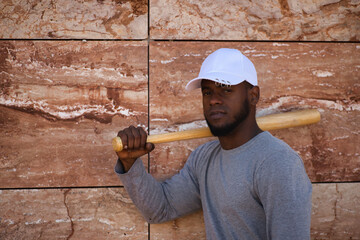 African american in white cap with baseball bat resting on his shoulder against a stone background.