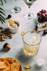 Whiskey is poured into a glass with ice. there are chips, glasses and other snacks nearby