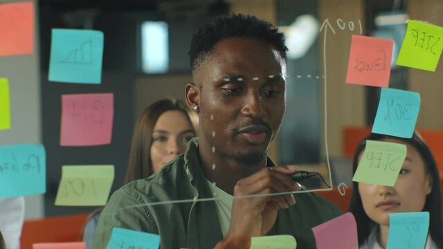 Young afro american man in casualwear drawing business chart on glass board with marker and speaking to team of coworkers during informal meeting in office.