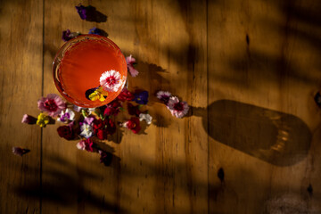 Top view of Aperitif cocktail in vintage glass with small flowers as decoration, on the side of wooden table. Subtle sunset light and shadows.