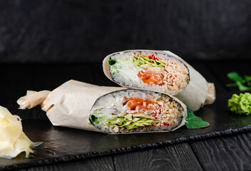 Salmon Sushi Burrito on black slate stone plate served with ginger slices and wasabi, wooden background.
