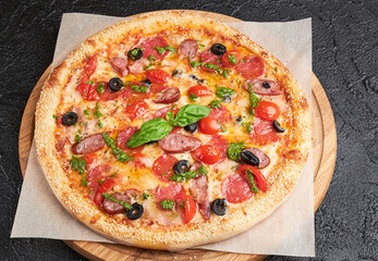 Whole Italian pizza with meat served with fresh basil leaf on parchment paper. Wooden board on dark background. Hot pizza with tomatoes, bacon, smoked sausages, chicken, olives and pesto sauce 
