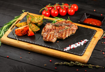 Well done Pork steak served with grilled potatoes on board with ingredients on dark background. Serving size of meat dish
