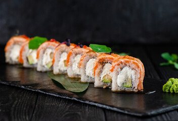Japanese sushi roll on Black slate stone plate on wooden background. sushi pieces with Philadelphia cream cheese, avocado wrapped in rice with burned salmon Tataki fillet slices on top with greens
