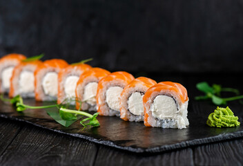 Japanese sushi roll on Black slate stone plate on black wooden background. Asian sushi roll pieces with Philadelphia cream cheese wrapped in rice with salmon fillet slices on top served with greens

