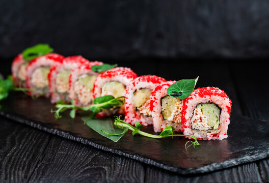 Japanese Sushi Roll On Black Slate Stone Plate On Wooden Background. Sushi Pieces With Cucumber, Crab Meat Sticks Wrapped In Rice With Red Flying Fish Roe On Top Served With Sprouts Of Fresh Herbs
