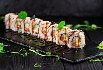 Japanese sushi roll on Black slate stone plate on wooden background. sushi pieces with salmon, cucumber wrapped in rice with eel slices on top served with unagi sauce, sprouts of fresh herbs
