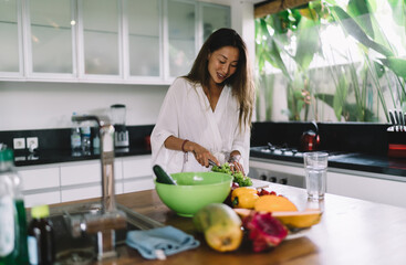 Smiling young Asian lady cutting lettuce while preparing dish in kitchen
