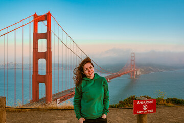A young woman in a green hoodie stands on a hill overlooking the Golden Gate Bridge during sunset,...