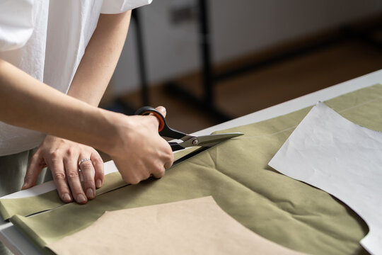 Cutting patterns in tailoring workshop concept: closeup of female hand using scissors to cut fabric. Seamstress at work. Tailor prepare material for sewing clothes. Fashion designer workplace studio