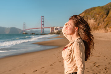 Beautiful young woman with long hair walks on the beach overlooking the Golden Gate Bridge in San...