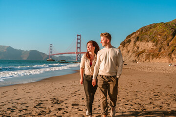 A young man and a woman take a romantic walk on the beach overlooking the Golden Gate Bridge at...
