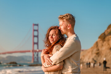 A young couple in love, a man and a woman, embrace on the beach in front of the Golden Gate Bridge...