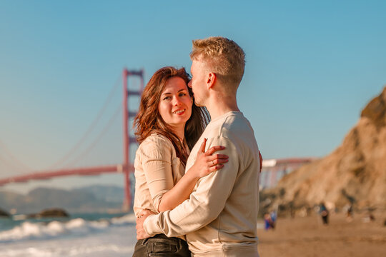A young couple in love, a man and a woman, embrace on the beach in front of the Golden Gate Bridge in San Francisco