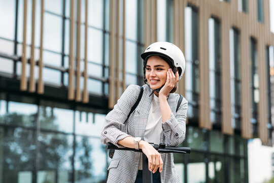 Positive Woman Leaning On Handlebar Of Electric Push Scooter. Pretty Female In Helmet Looking Away While Standing In The City.