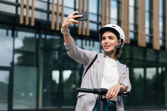 Young beautiful woman in a white cycling helmet taking selfie while standing on an electric push scooter in the city