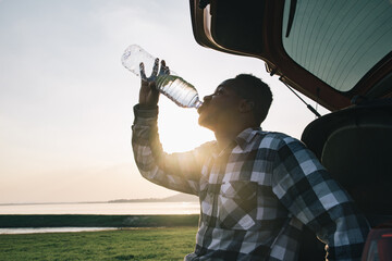 African American man tourists relax in front of their cars and drink thirsty water from bottle with sunlight. In evening atmosphere of the dam with mountains and good weather for relaxing and camping