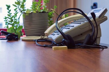 Headphones with microphone, telephone set and houseplant on the surface of the office table. Telephone consultations