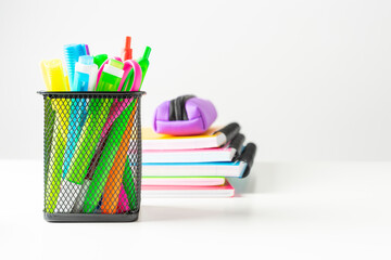 Group of markers, colored pens, scissors in a metal basket next to several colored notebooks on a white desk. Back to school concept