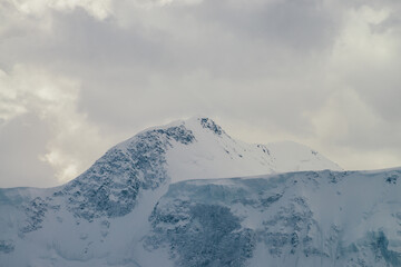 Atmospheric highland landscape with high snowy mountain wall under cloudy sky. Dramatic scenery with snow-covered great mountain top in overcast weather. Sunlight through clouds above snow pinnacle.
