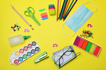 On a yellow background, there are school supplies, an eraser, scissors, pencils, a protective medical mask, paints, glasses, a stapler. Back to school.