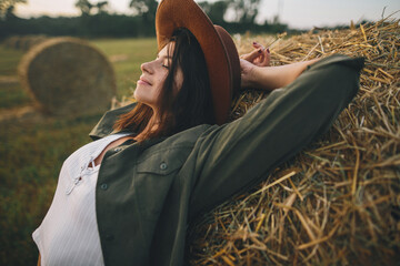 Beautiful stylish woman in hat relaxing on haystack in summer evening field. Portrait of attractive...
