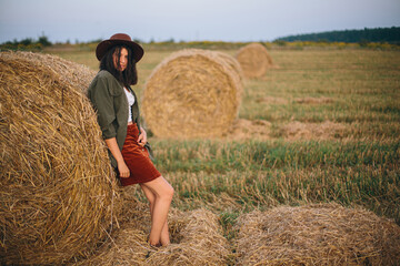 Naklejka na ściany i meble Stylish woman in hat standing at hay bale in summer evening in field. Atmospheric tranquil moment. Young fashionable female relaxing at haystack, summer vacation in countryside. Rural slow life