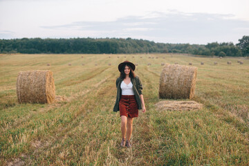 Naklejka na ściany i meble Beautiful stylish woman in hat walking at hay bales in summer evening field. Happy young female relaxing at haystacks, vacation in countryside. Tranquility, countryside slow life