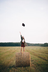 Fototapeta na wymiar Beautiful carefree woman throw up hat in the sky while standing on haystack in evening summer field. Happiness. Young happy female having fun on hay bales in countryside. Atmospheric moment