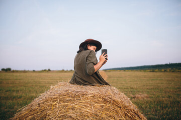 Naklejka na ściany i meble Beautiful stylish woman sitting on haystack and taking photo of sunset on phone in summer field. Young female holding smartphone and capturing evening in warm sunshine. Atmospheric moment