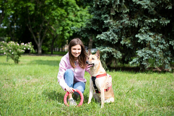 Naklejka na ściany i meble Portrait of a young cheerful woman sitting in the park on the green grass with a dog from the shelter.