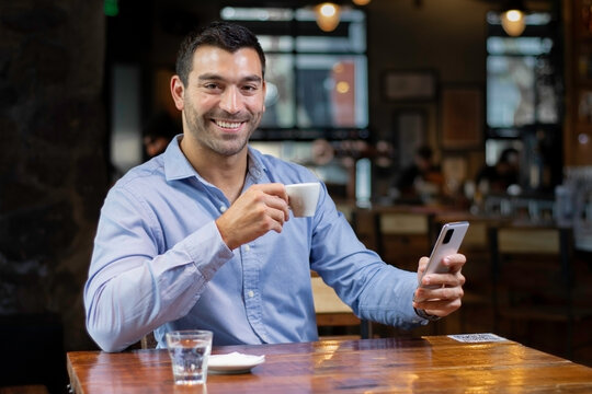 Hombre joven enviando mensaje por el movil mientras toma cafe en una cafeteria moderna