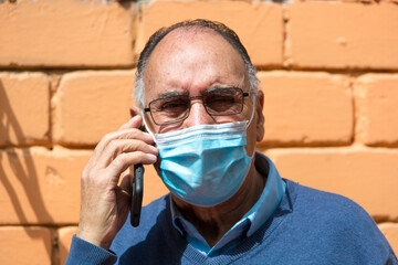 Portrait od an old senior man with glasses and medical face mask talking on the phone outside with a orange brick wall in the background. Scene of COVID or Coronavirus pandemic.