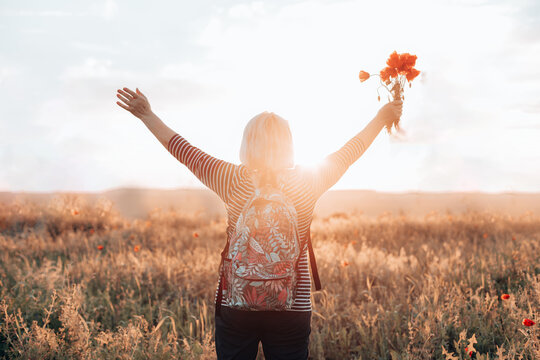 Adult woman tourist with backpack and poppy flower bouquet at sunset in the field. summer vacation. Happy girl with blond hair enjoying the sun. Horizontal banner