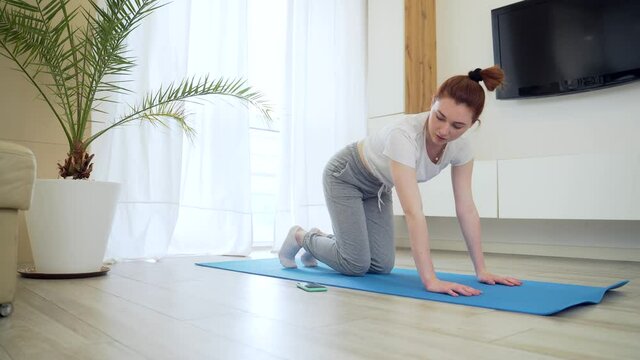 young redhead girl woman in casual clothes doing physical exercise using mobile phone app with workout. female on mat stretching Online tutorial of yoga, aerobics, fitness pilates at home living room