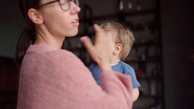 Young Mother Holding Baby Son In Her Arms At Home. Anxious Baby Hitting Her Face. Loving Mom Soothing And Calming Upset Kid, Embracing Him Gently, Stroking And Explaining Him Things