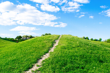 Fototapeta na wymiar Small hiking trail, path climbing up the mountain, hill, mound with green grass against bright blue sky with clouds
