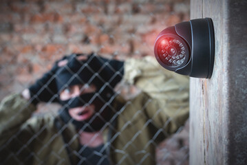 Security camera on the wall close up and confused robber in the mask behind the fence concept...