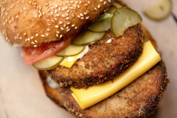 Double meat burger. Closeup of Bun with two fried cutlets, cheese, salted cucumber and tomato. Super hearty snack. Horizontal format. Soft focus.