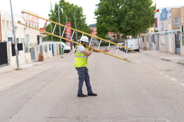 telecommunications technician crossing the street with ladder over his left shoulder, wearing work clothes and apparently tired.